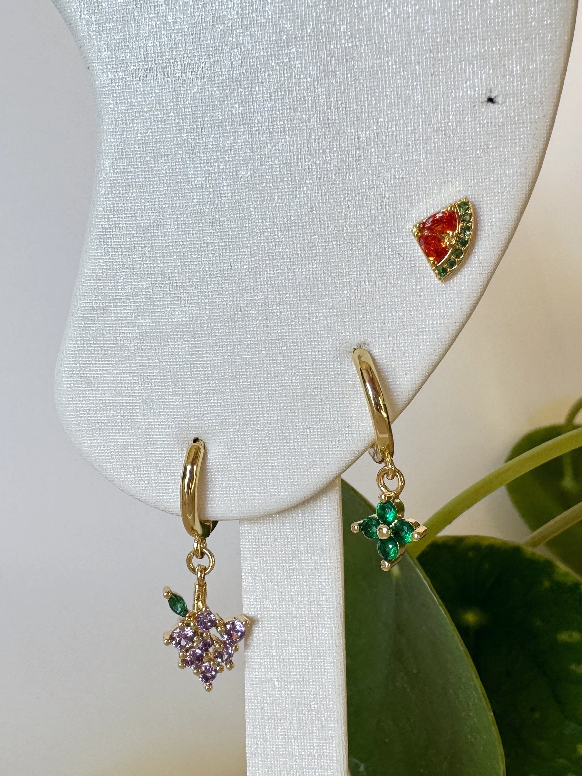 Gold earrings with green and purple gemstones on a white stand with leaves in the background