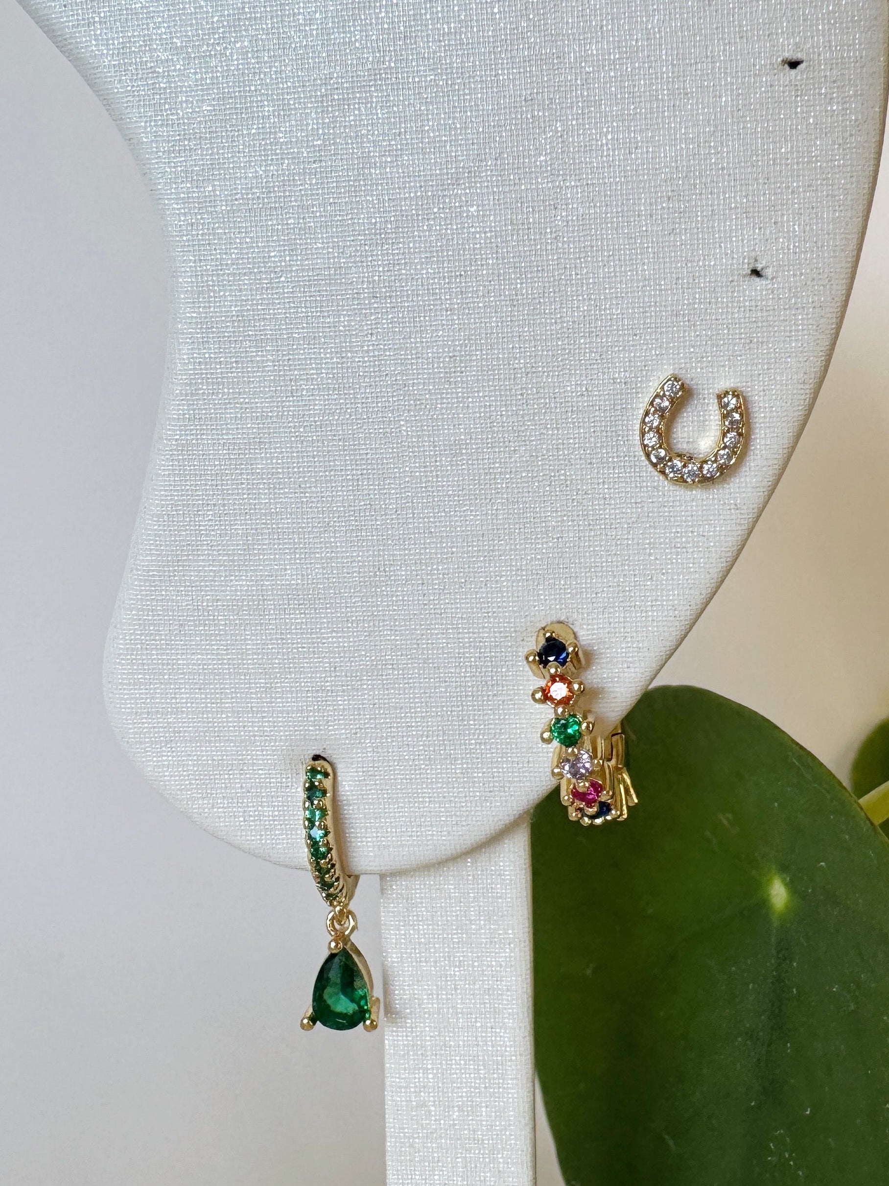 Three pairs of green and gold earrings including horseshoe studs on a white stand with a plant in the background