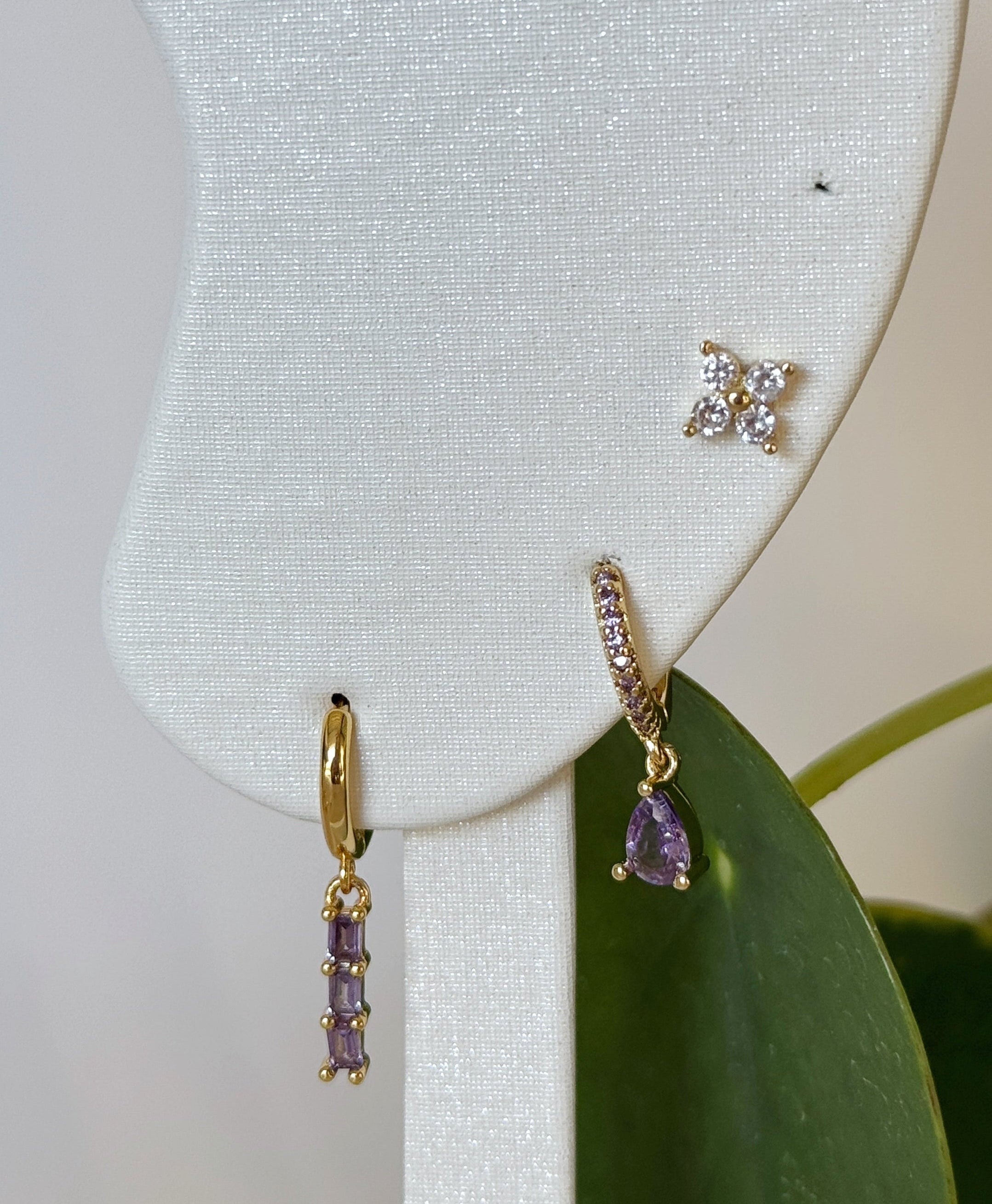 Gold earrings with purple gemstones on a white earring stand with green leaves in the background