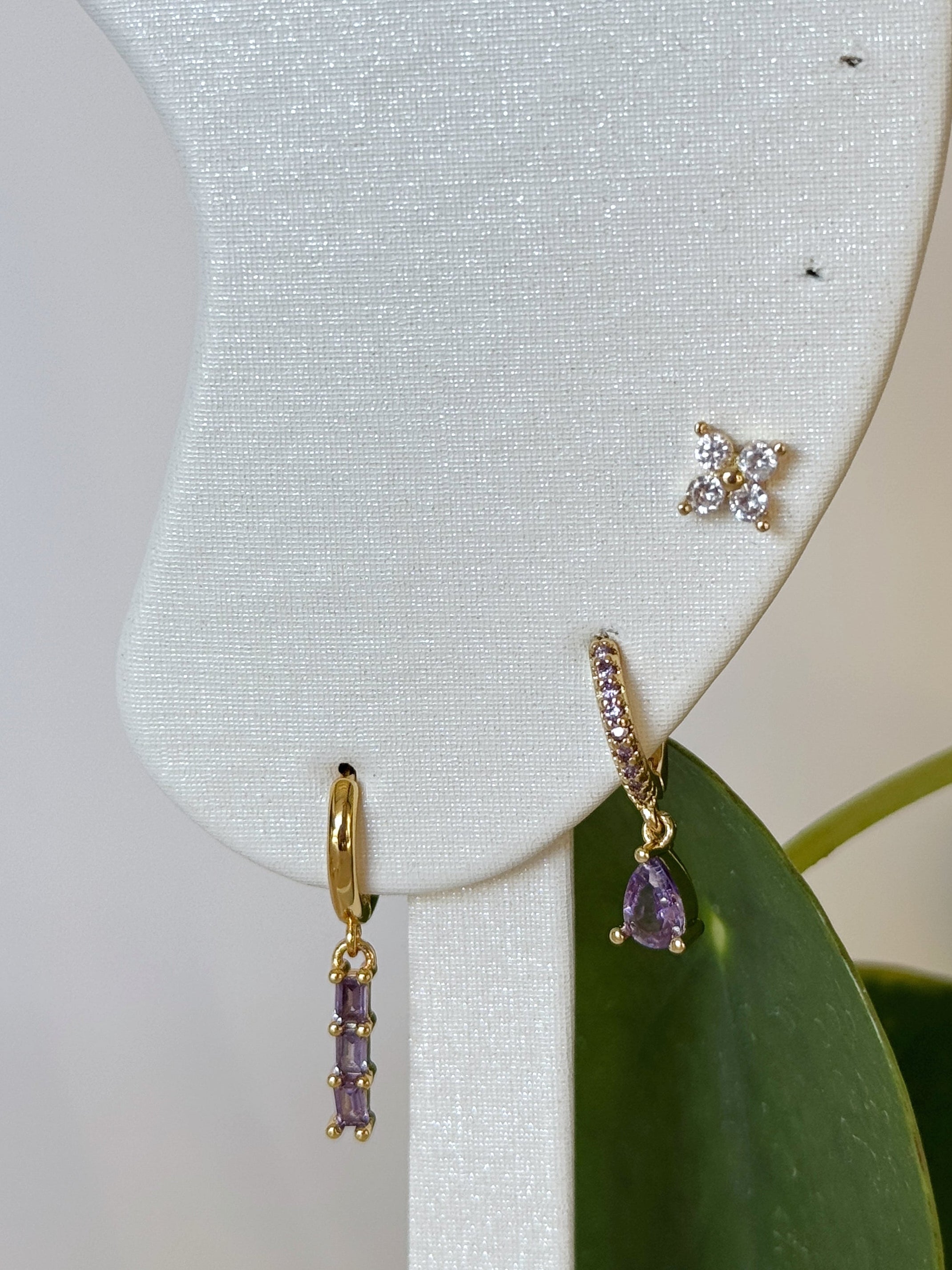 Gold and lavender stainless steel earrings with gemstones on a white earring stand with green leaves in the background