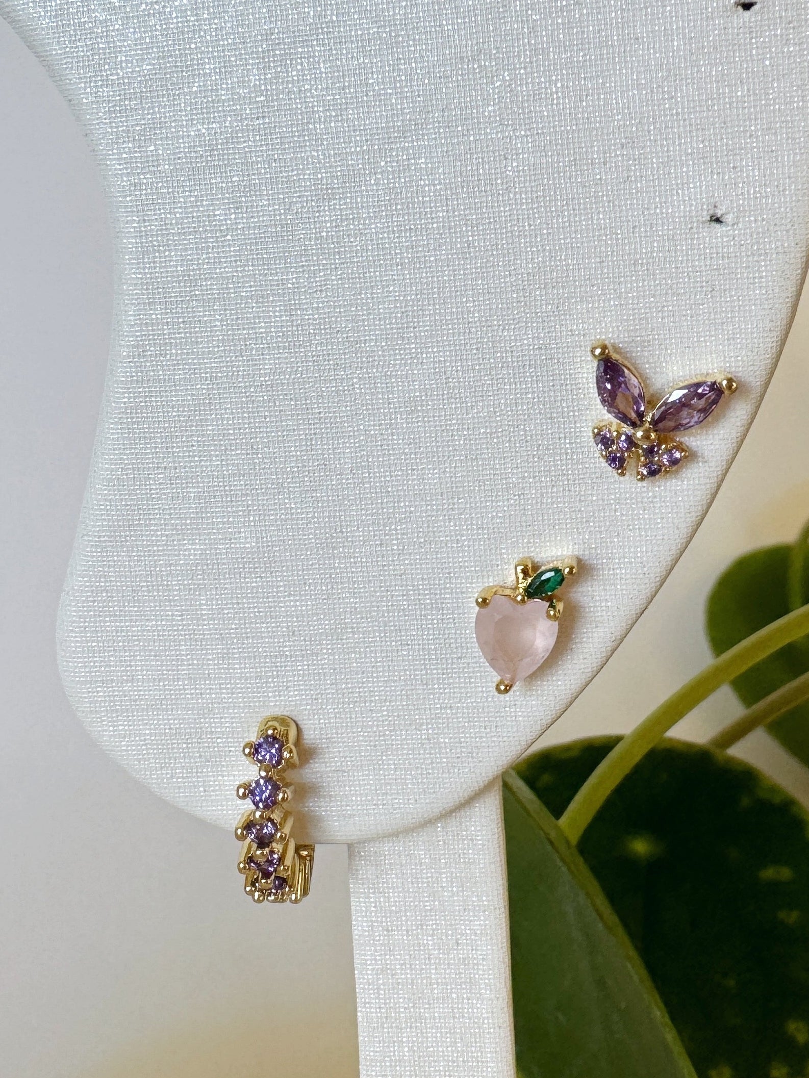 Three pairs of waterproof earrings on a white stand with green leaves in the background, purple butterfly and pink peach + lavender hoop