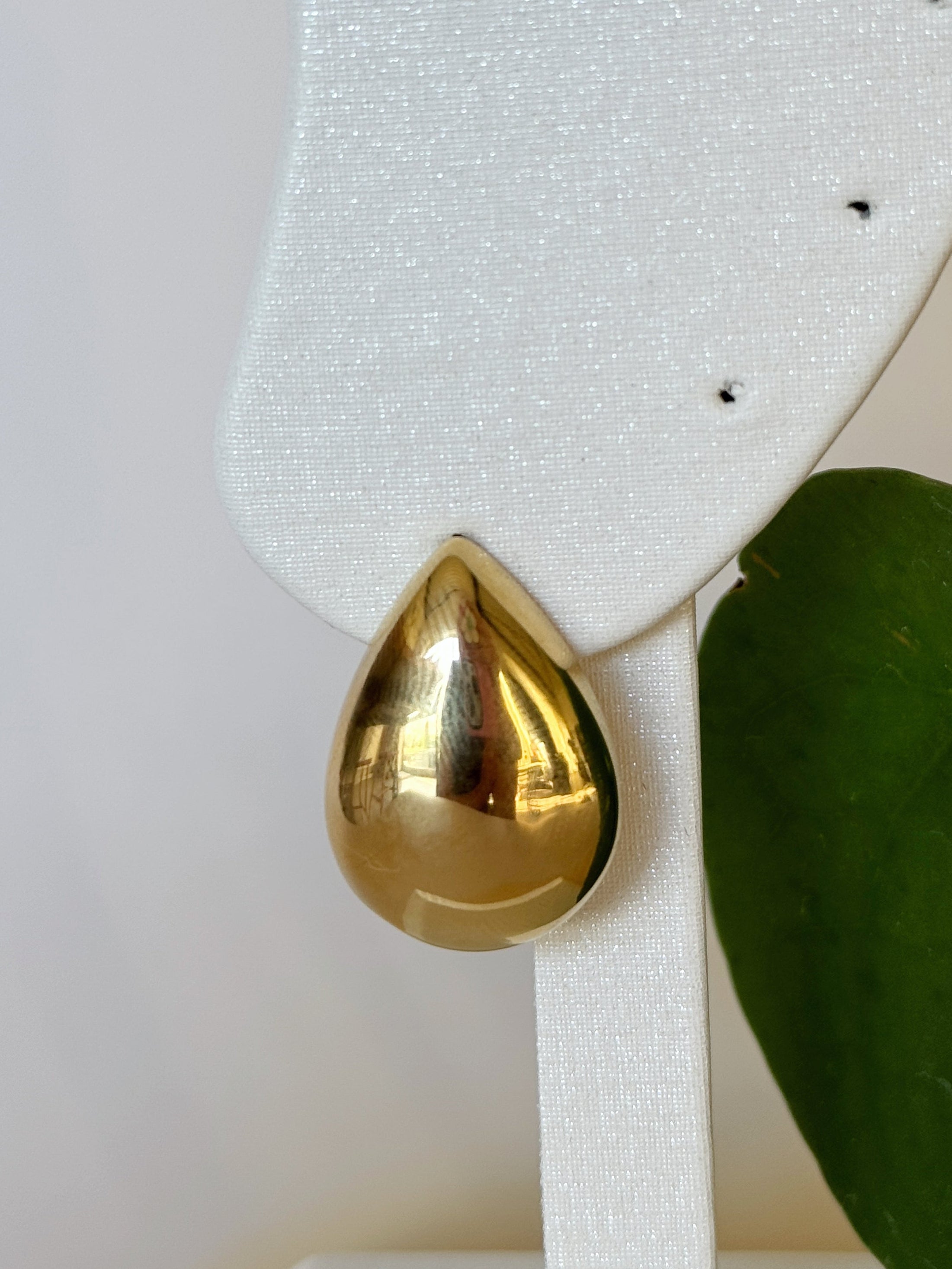 Gold teardrop earring on a white stand with a green leaf in the background