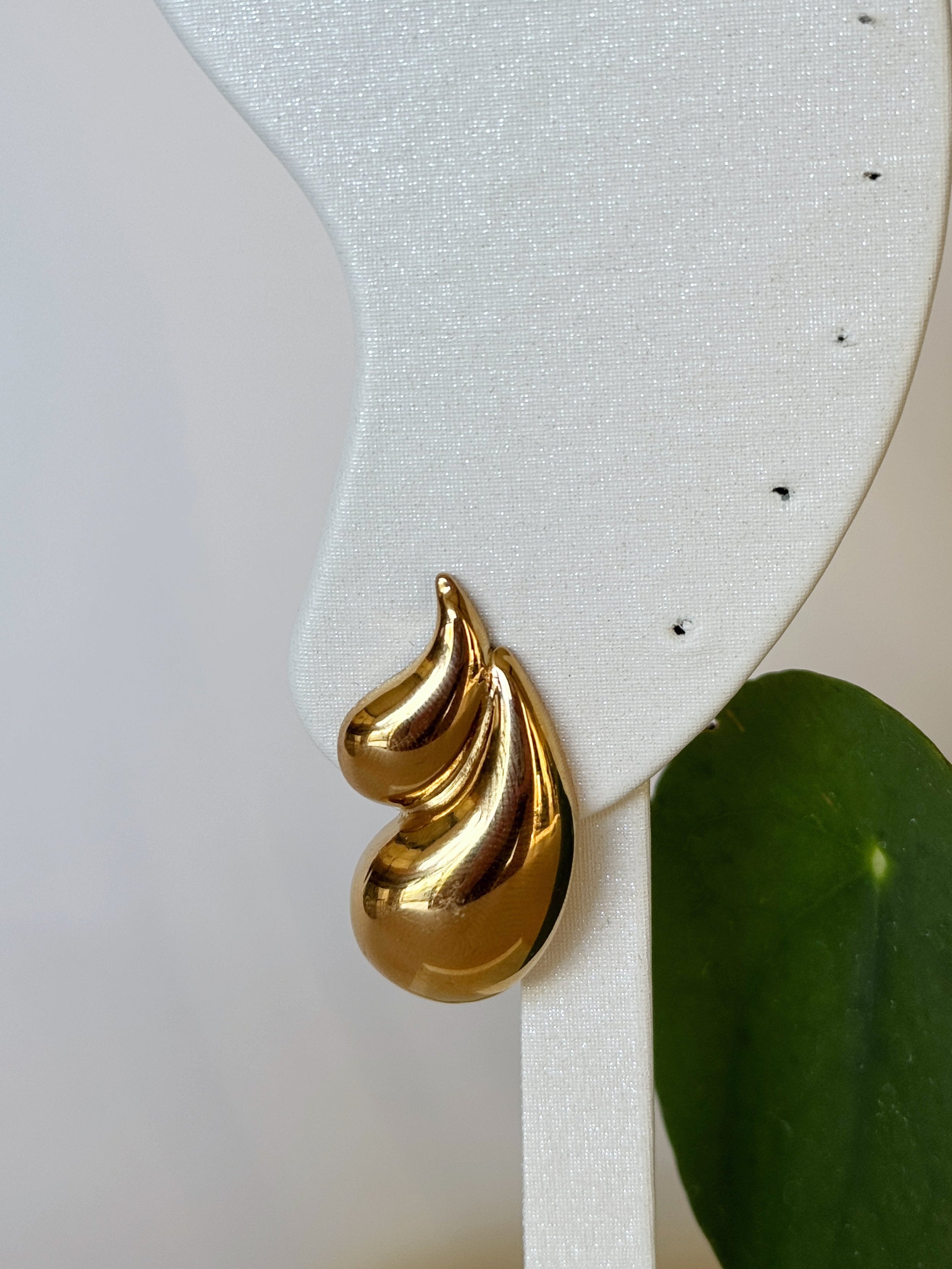 Gold spiral teardrop earring on a white stand with a green leaf in the background