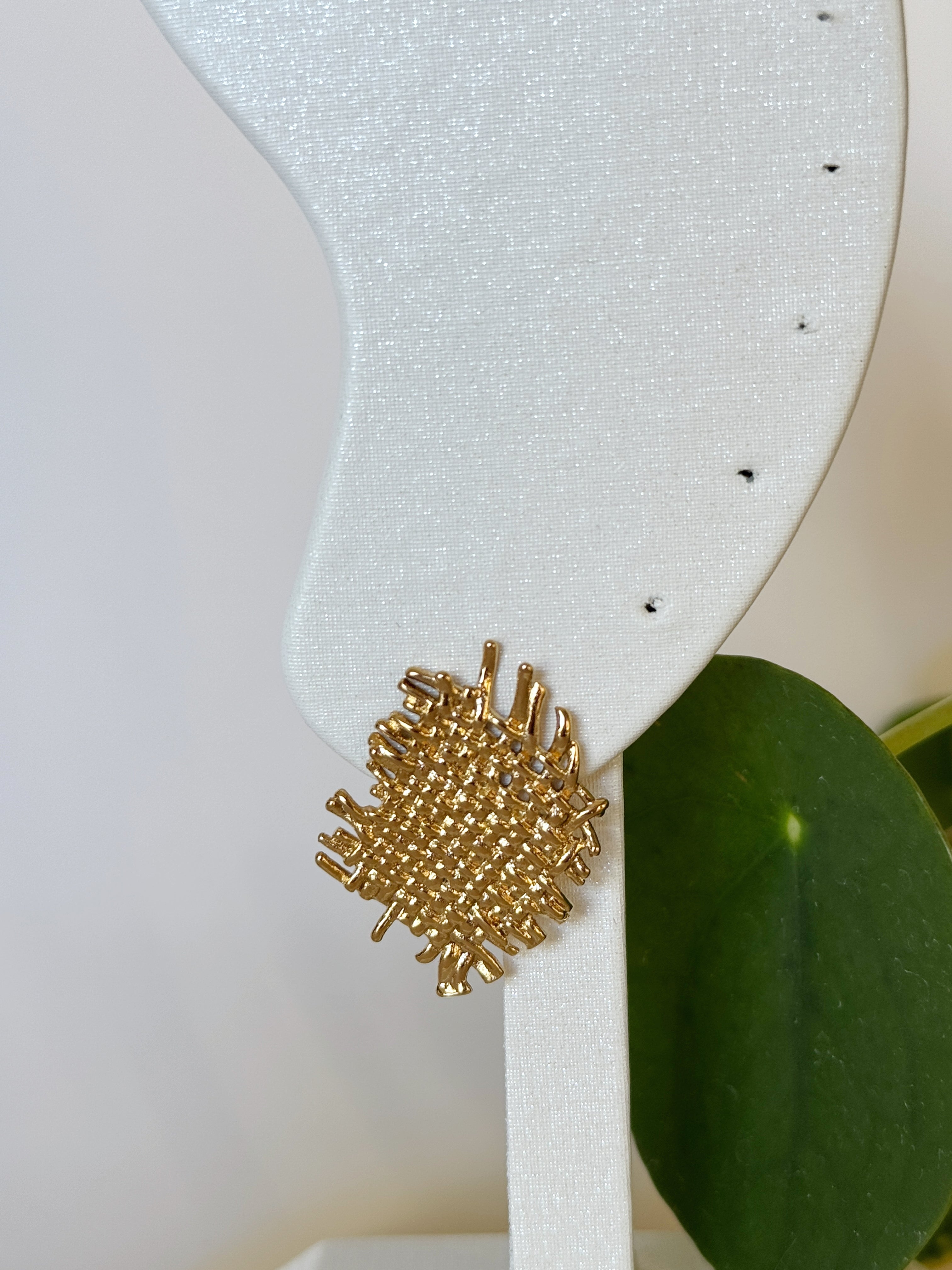 Gold textured earring on a white card with green leaves in the background