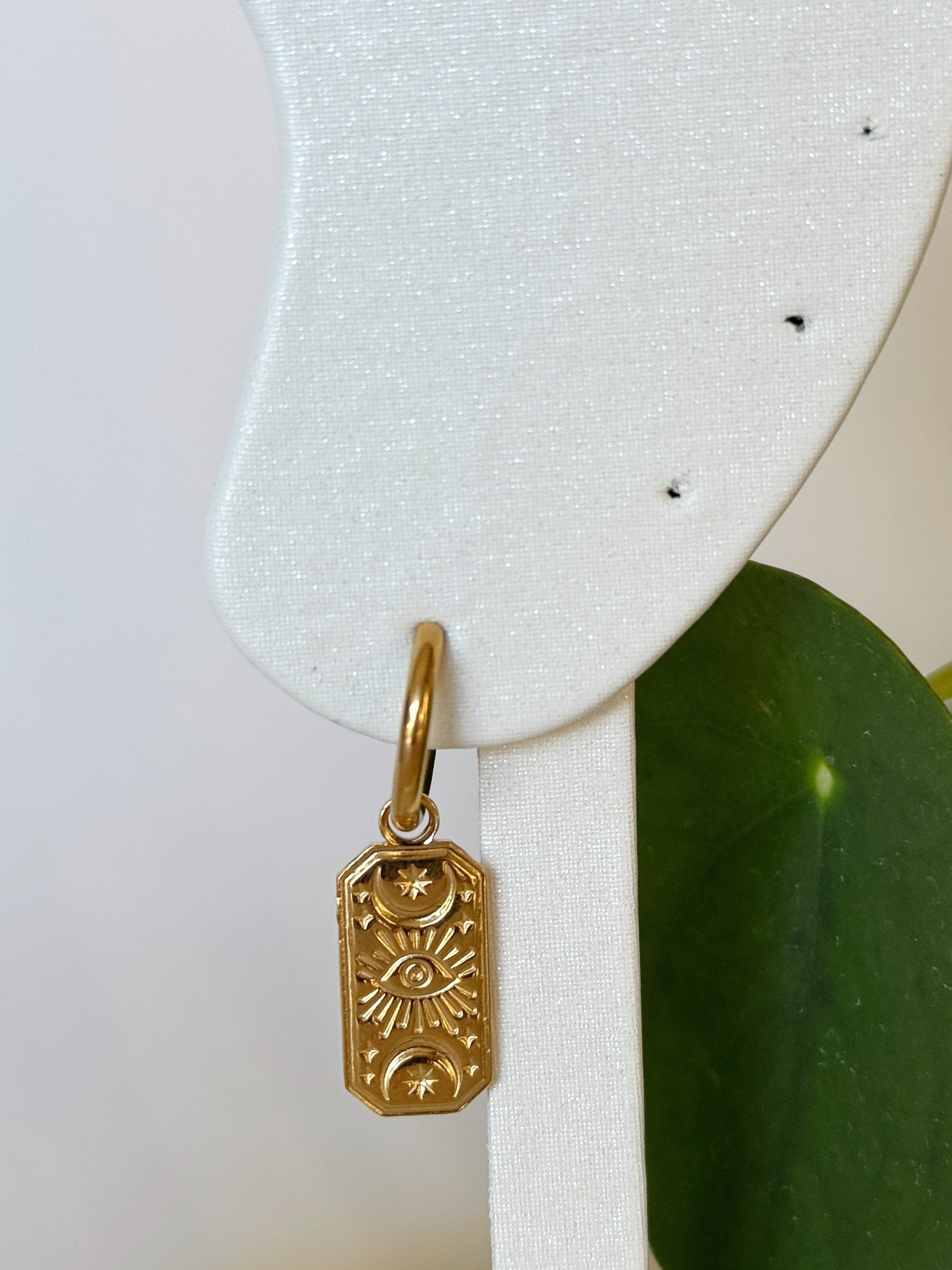 Gold stainless steel evil eye pendant earring on a white earring stand with green leaves in the background