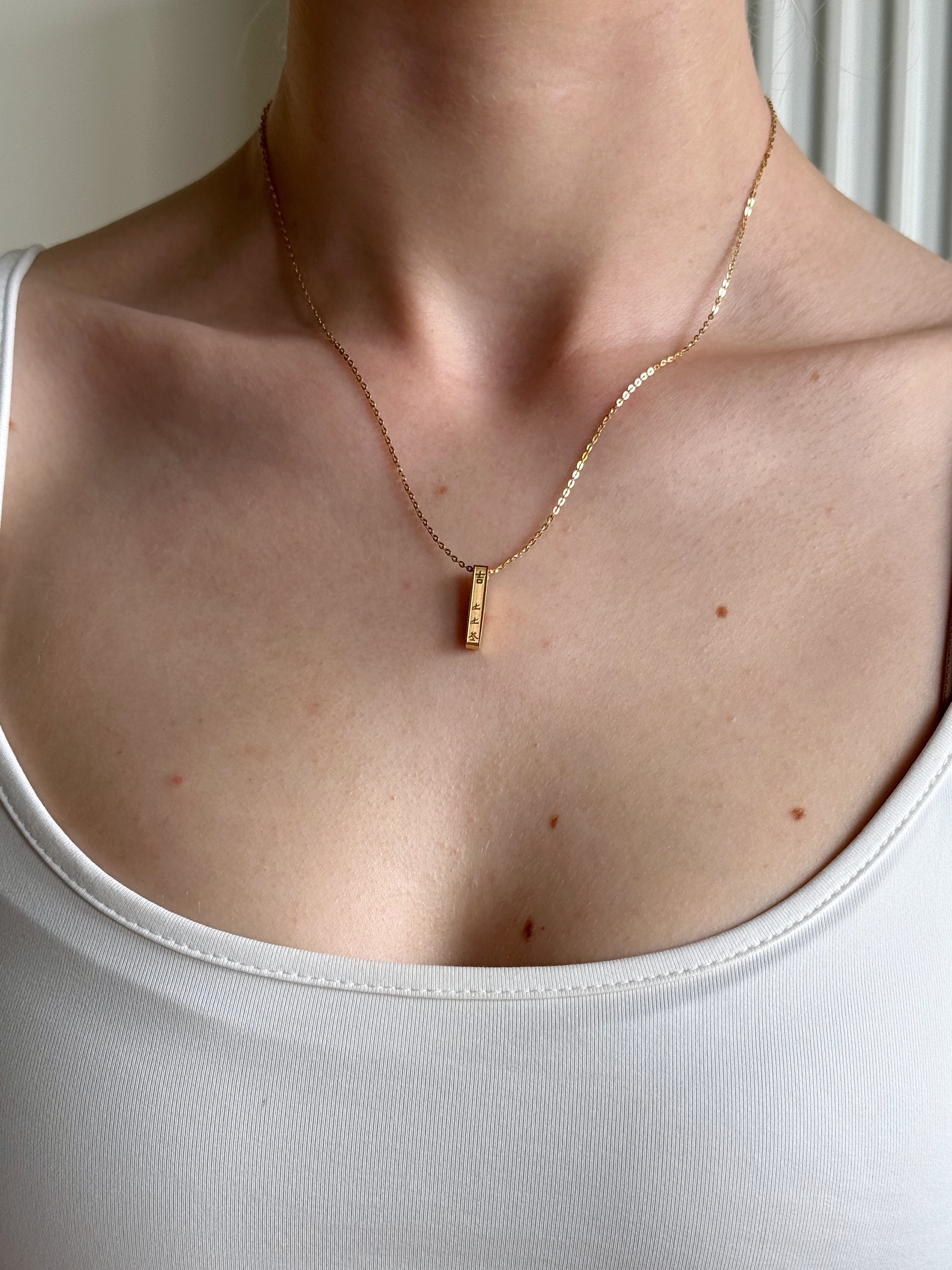 Gold necklace with a bar pendant with  a Chinese good luck message on a person wearing a white tank top.